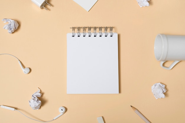 Blank white notepad surrounded by empty cup; crumpled paper; pencil; earphone over beige backdrop Free Photo