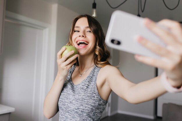 りんごで自分の写真を撮る陽気な白人の女の子 自撮りにスマートフォンを使って笑う愛らしい女性 無料の写真