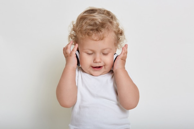 白いスペースにヘッドフォンで金髪の縮れ毛の赤ちゃん 目を閉じて音楽を楽しんでいる子供 プレミアム写真