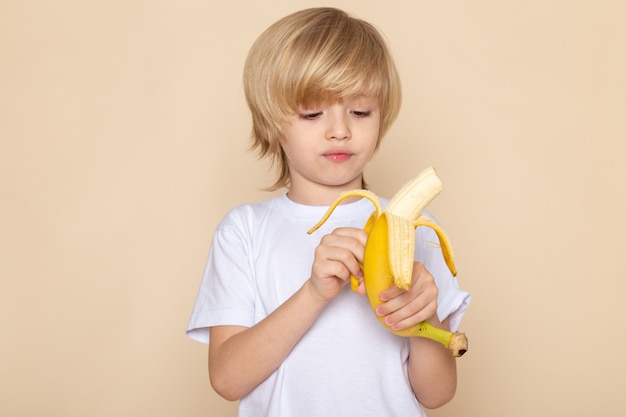 金髪の少年かわいい愛らしいピンクの白いtシャツでバナナを皮をむく 無料の写真