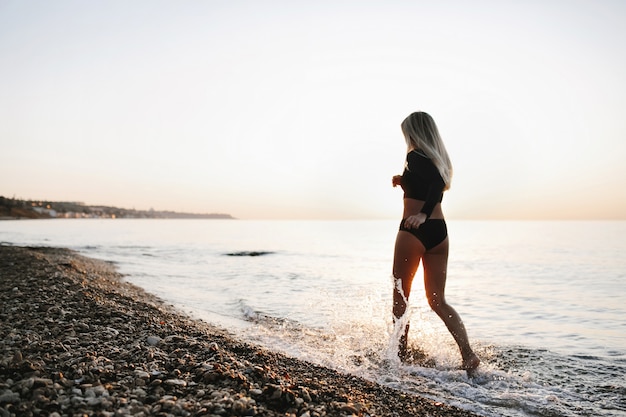 夕日に海の海岸で黒い水着のブロンドの女の子が実行されています 無料の写真