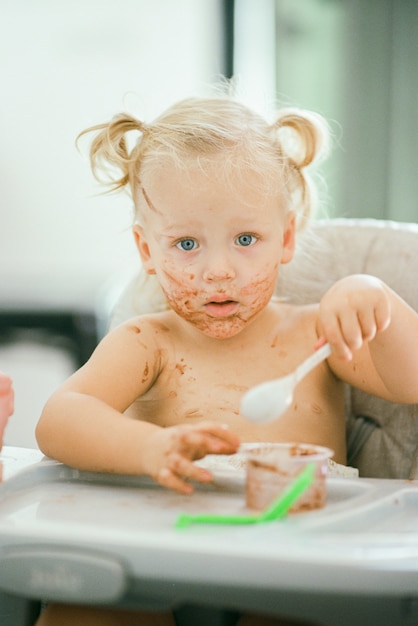 ハイチェアに座って食事をするときの汚い顔で金髪の髪の赤ちゃん女の子 プレミアム写真