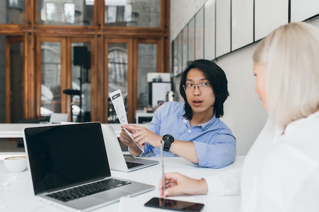 Free Photo Blonde Secretary Sitting With Phone Beside Laptop With Black Screen And Listening Asian Young Man In Glasses Brunette Chinese Office Worker Talking With Female Manager In White Blouse