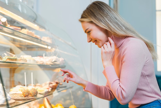 Blonde Young Woman Looking Through Cake Display Cabinet In Cafe
