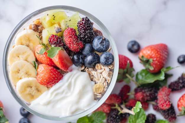 Bowl of oat granola with yogurt, fresh blueberries, mulberry, strawberries, kiwi, banana, mint and nuts board for healthy breakfast, top view, copy space, flat lay. vegetarian food concept. Free Photo