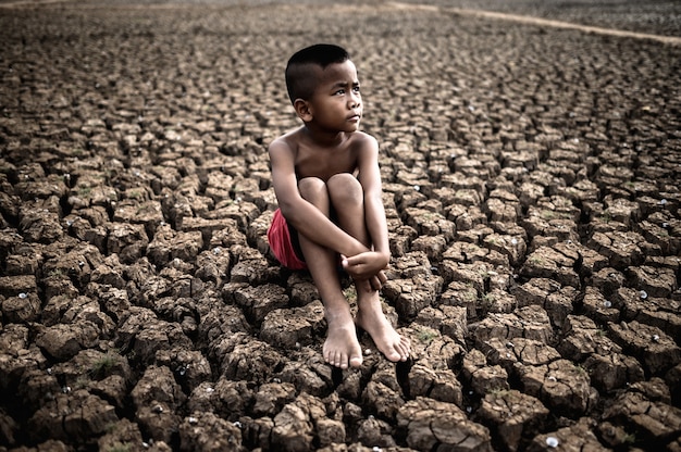The boy sit hugging their knees bent and looking at the sky to ask for rain on dry soil. Free Photo