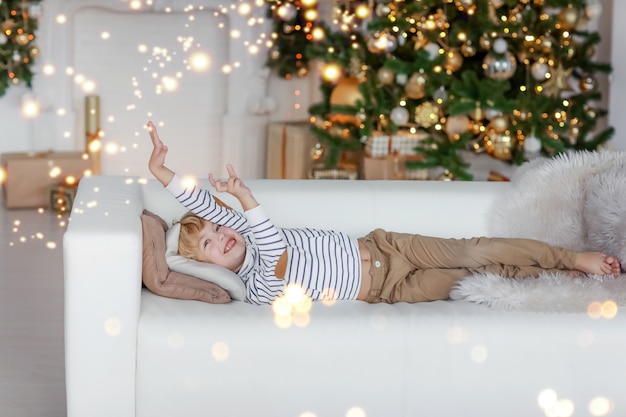 Download Premium Photo A Boy With Blond Hair Against The Background Of A Christmas Tree Lies On A Sofa And Dreams Of The Best Santa Claus Gift PSD Mockup Templates