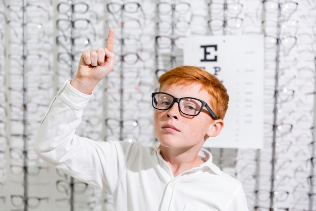 Boy with spectacle pointing at upward direction standing against eyeglasses display background Free Photo