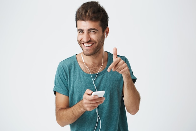 Free Photo Brightly Smiling Unshaved Spanish Guy In Blue T Shirt Holding Smartphone Listening Music With Headphones Laughing And Gesticulating Positive Human Facial Expressions And Emotions