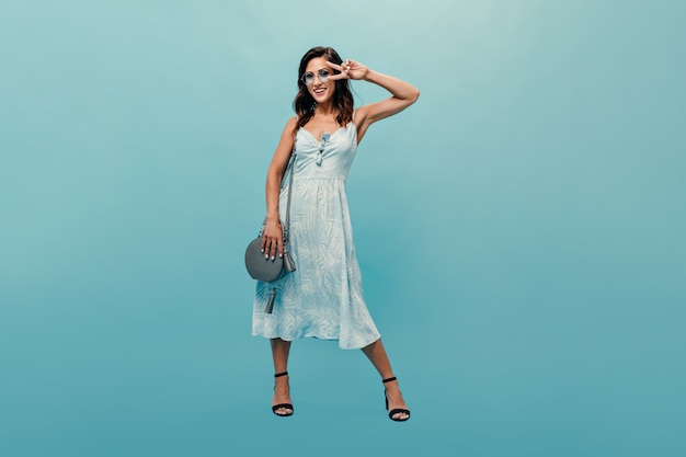Free Photo Brunette Woman In Dress And Stylish Glasses Shows Peace Sign On Blue Background