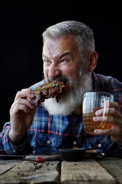 ひげを持つ残忍な白髪の成人男性はマスタードステーキを食べるし ビールを飲む 食事 休日の概念 祭り オクトーバーフェストに招待します プレミアム写真