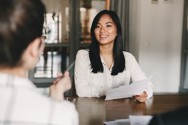 Business, career and placement concept - joyful asian woman smiling and holding resume, while sitting in front of directors during corporate meeting or job interview Premium Photo