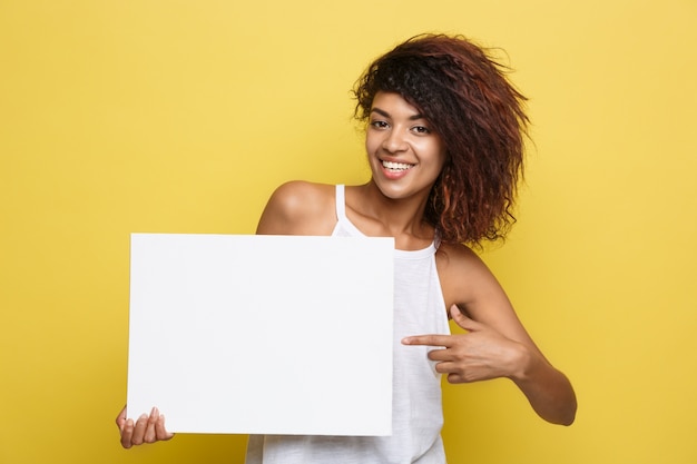 Business concept - close up portrait young beautiful attractive african american smiling showing plain white blank sign. yellow pastel studio background. copy space. Free Photo