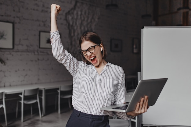 Business woman with laptop in hand is happy with success. portrait of woman in glasses and striped blouse enthusiastically screaming and making winning gesture. Free Photo