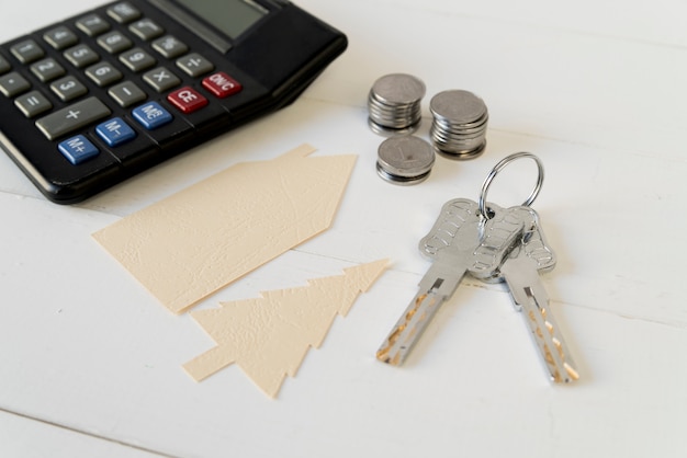 Calculator; coins stack; keys with house and tree paper cutout on white wooden table Free Photo