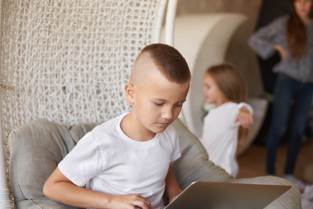 Candid shot of serious concentrated schoolboy with stylish haircut sitting on chair at home, using laptop computer while doing homework, his family talking in background. childhood and technology Free Photo