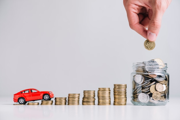 Car driving over increasing stacked coins near person's hand putting coin in glass jar Free Photo