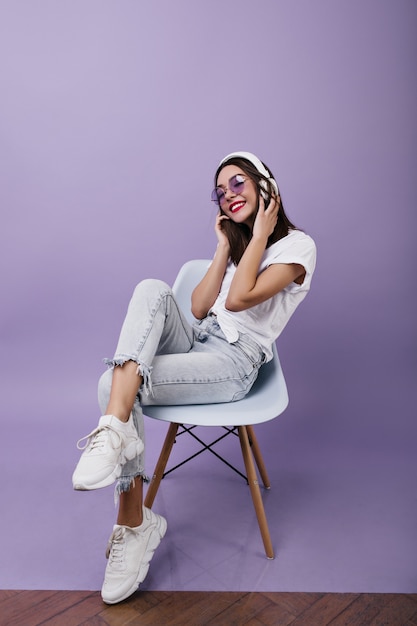 椅子に座って音楽を聴いている流行のスニーカーでのんきな若い女性 ヘッドフォンポーズでファッショナブルなブルネットの女性モデル 無料の写真