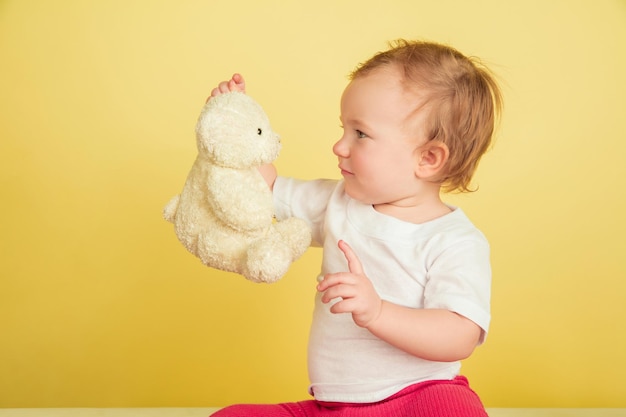 Caucasian little girl, children isolated on yellow studio background. portrait of cute and adorable kid, baby playing with teddy bear. Free Photo