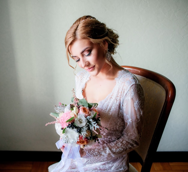 Charming blonde bride in sexy white robe sits with wedding bouquet on the chair Free Photo