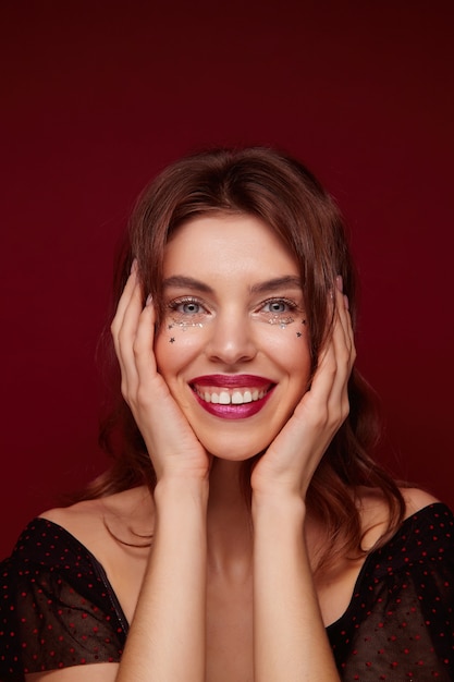 Free Photo Charming Blue Eyed Young Lady With Brown Wavy Hair Wearing Silver Stars On Face And Holding Palms On Her Cheeks Smiling Cheerfully To Camera While Posing Against Claret Background