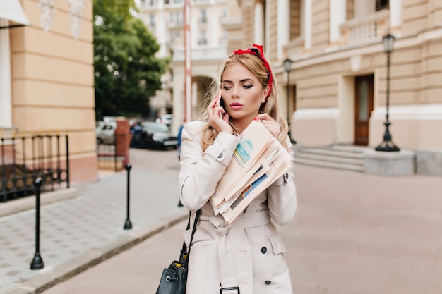 Free Photo Charming Business Lady With Elegant Make Up And Blonde Hair Is Rushing To Work Outdoor Portrait Of Young Woman In Beige Coat Holding Newspaper And Talking On Phone