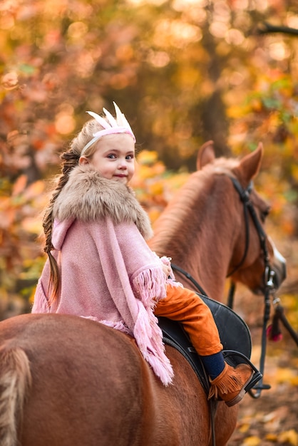 Charming little girl dressed like a princess rides a horse around the autumn forest Free Photo