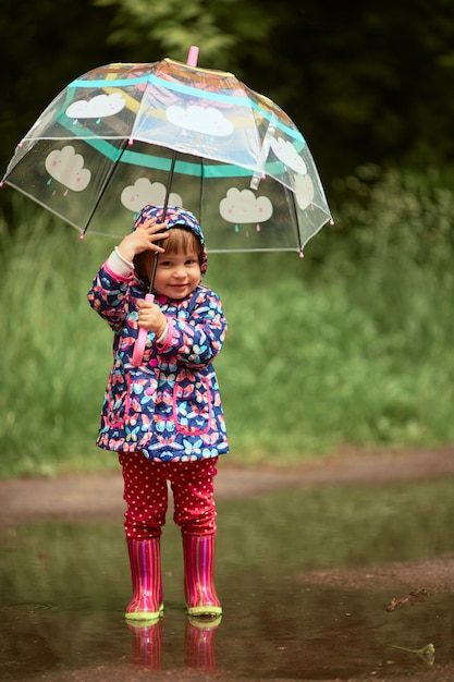 Charming little girl with umbrella has fun standing in gumboots in the pool after rain Free Photo