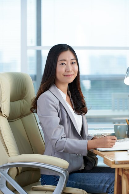 Cheerful asian woman sitting at desk in office, writing and posing for