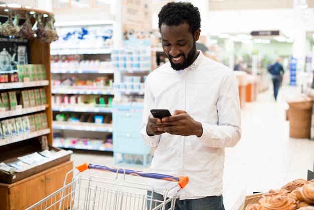 Cheerful black man typing on cellphone in grocery store Free Photo