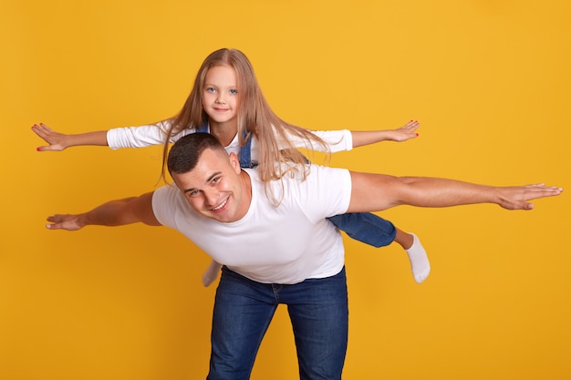 陽気な幸せな男が彼の素敵な娘と一緒に飛行機のように飛ぶことを想像して 黄色に分離されたポーズをとります 最高の父親 一体感 家族の概念とのショット 幸せな瞬間 無料の写真