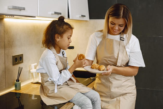 Cheerful mother and little daughter eating fresh baked cookies in kitchen, enjoying homemade pastry, wearing aprons and smiling to each other, having fun at home. Free Photo