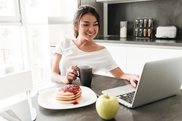 Free Photo Cheerful Young Woman Sitting At The Kitchen Using Laptop