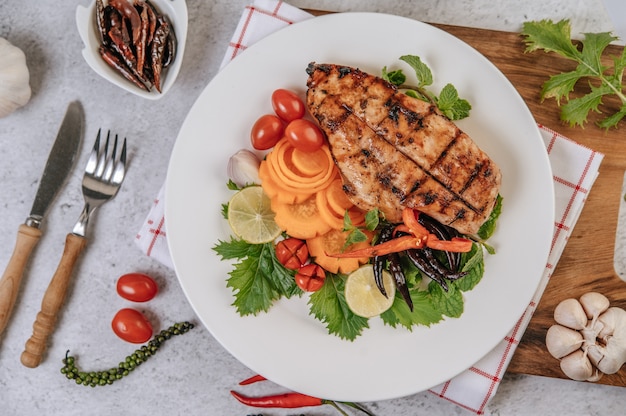 Chicken steak with lemon, tomato, chili, and carrot on white plate. Free Photo