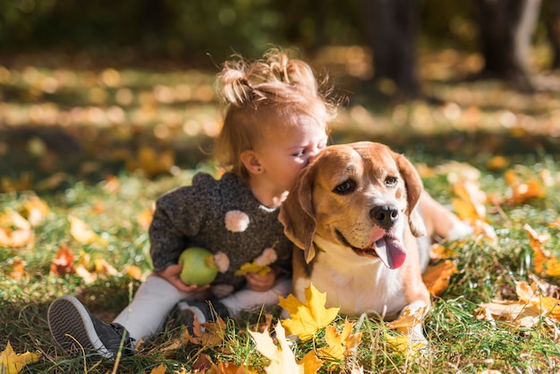 子供の女の子が森の芝生に座っている彼女の犬にキス 無料の写真