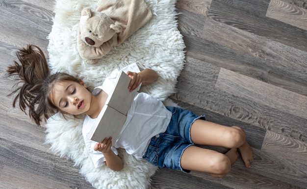The child reads a book lying on a cozy rug at home with his favorite toy teddy bear. Free Photo