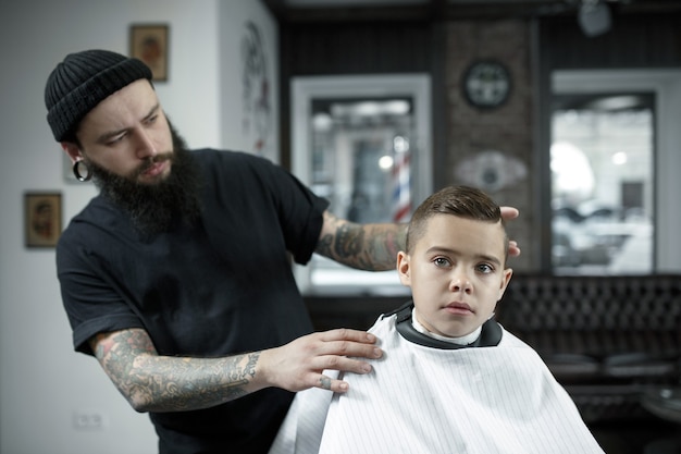 子供の美容師が暗い背景に小さな男の子をカットします 髪を切って満足しているかわいい幼児男の子 マスターの手には ひげをそるというタトゥーがあります 無料の写真