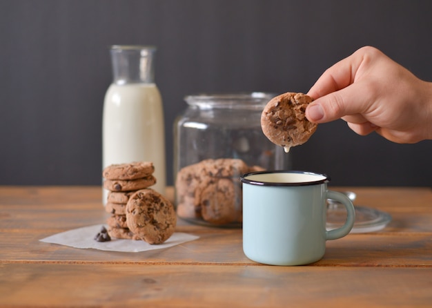 Chocolate chip cookies in glass jar with glass bottle of milk and turquoise enamel mug on wooden rustic table with men hand holding one cookie Premium Photo