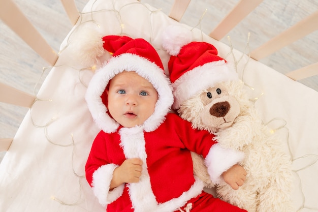 サンタクロースの帽子をかぶったおもちゃで自宅のベビーベッドに横たわっているサンタの衣装を着た赤ちゃんのクリスマスの写真 プレミアム写真