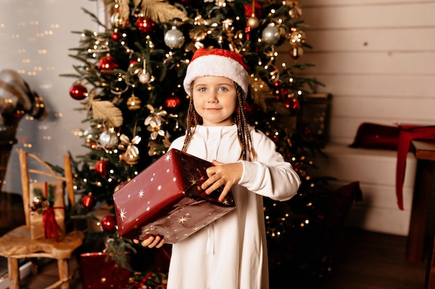 クリスマスの時期 クリスマスツリーの上にポーズをとってクリスマスプレゼントとクリスマス帽子の陽気な子供 無料の写真