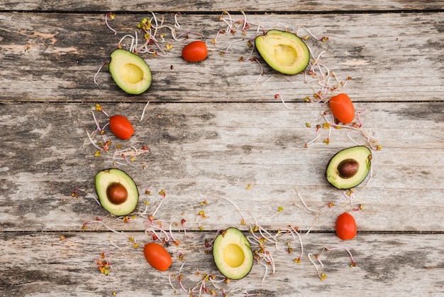 Circular frame made with halved avocado; tomatoes and sprouts on wooden desk Free Photo