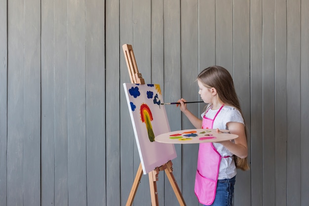 Close-up of a blonde girl painting with paint brush on easel standing against gray wooden wall Free Photo