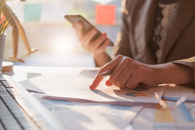 Close up of businesswoman or accountant hand holding pencil working on smart phone data report, accountancy document and laptop computer at office, business concept Premium Photo