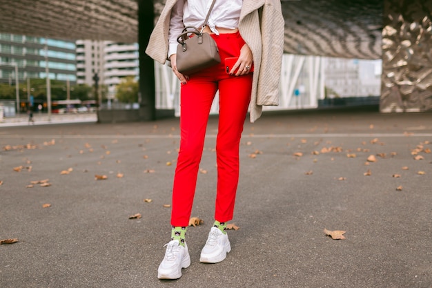 Close up fashion details , young woman wearing trendy red pants funny socks and ugly fashion sneakers, beige elegant coat, posing on the street near business centers, autumn time. Free Photo