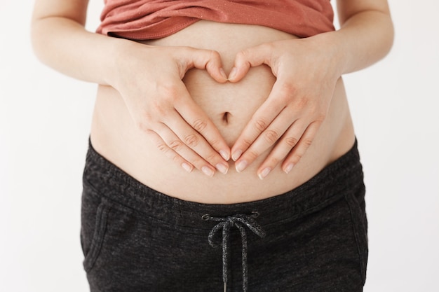 妊娠中の腹のクローズアップ カジュアルな服装の若い幸せな母は妊娠中の胃にハートのサインを作るに手を保持しています 無料の写真