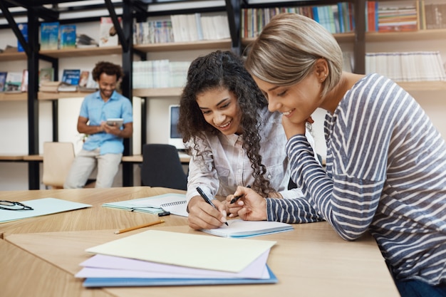 Close up of pair beautiful young multi-ethnic student girls doing homework together, writing essay for presentation, preparing for exams with good mood Free Photo