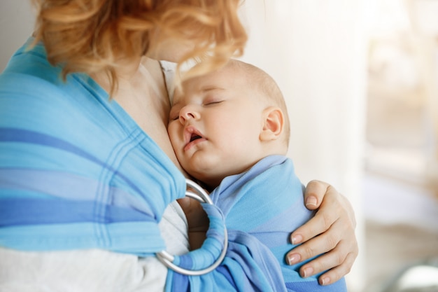 Close up portrait of innocent newborn boy having sweet dreams on mother chest in baby sling. mom looking at her child with love and tenderness. Free Photo