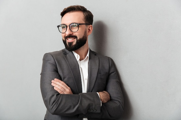 Close up portrait of pleased unshaved man in eyeglasses looking on camera with sincere smile, standing with arms folded isolated over gray Free Photo