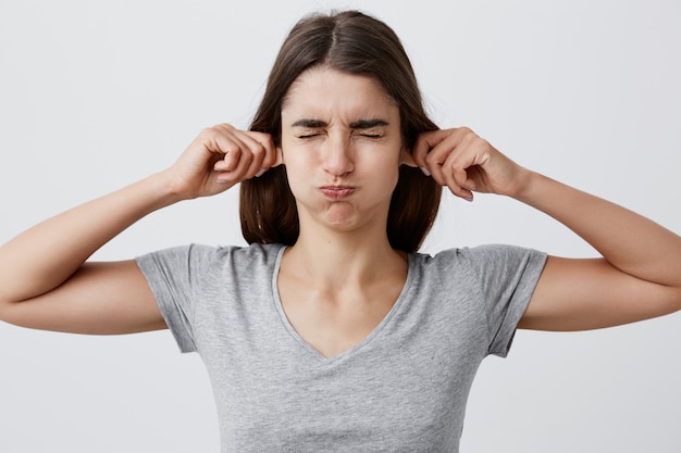 Free Photo Close Up Portrait Of Young Good Looking Funny Brunette Caucasian Girl With Long Hair In Casual Gray Shirt Holding Ears With Hands Closing Eyes Making Silly Faces To Make Little