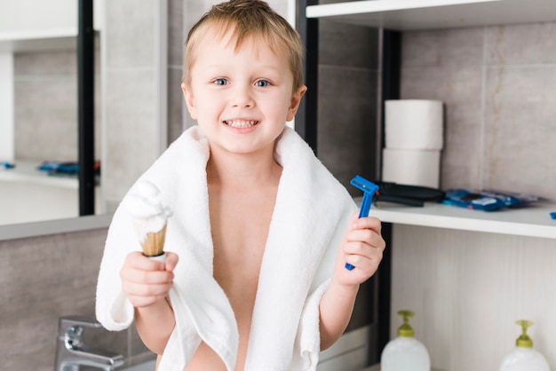 Close-up of a smiling boy standing in the bathroom holding shaving brush and blue razor in hand Free Photo